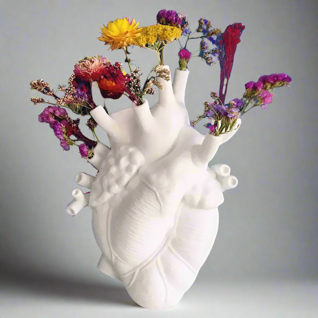 White anatomical heart-shaped vase with colorful flowers against a blue wall.