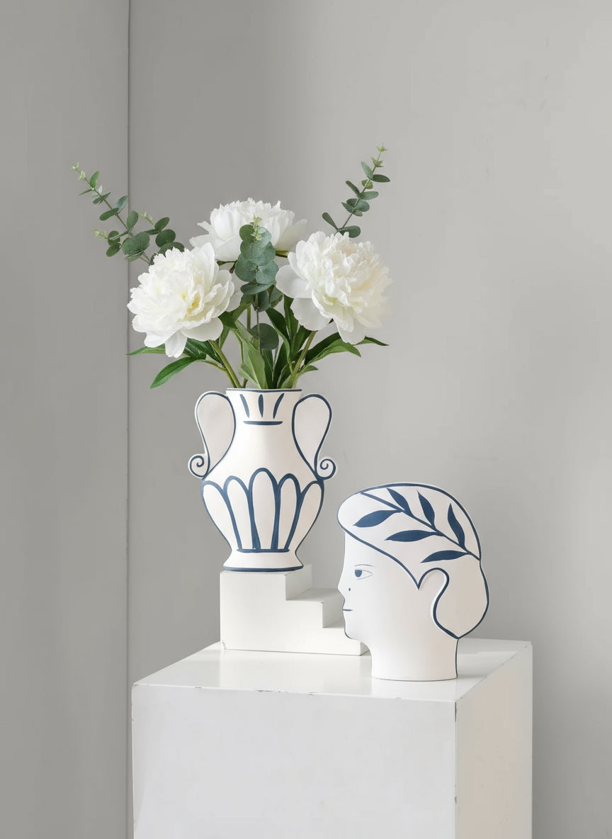 Decorative vase with flowers and a unique head-shaped container on a white surface against a beige wall.