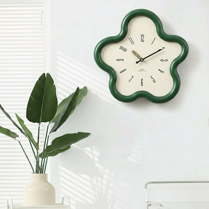 Flower-shaped wall clock with green frame on a white wall next to a plant.