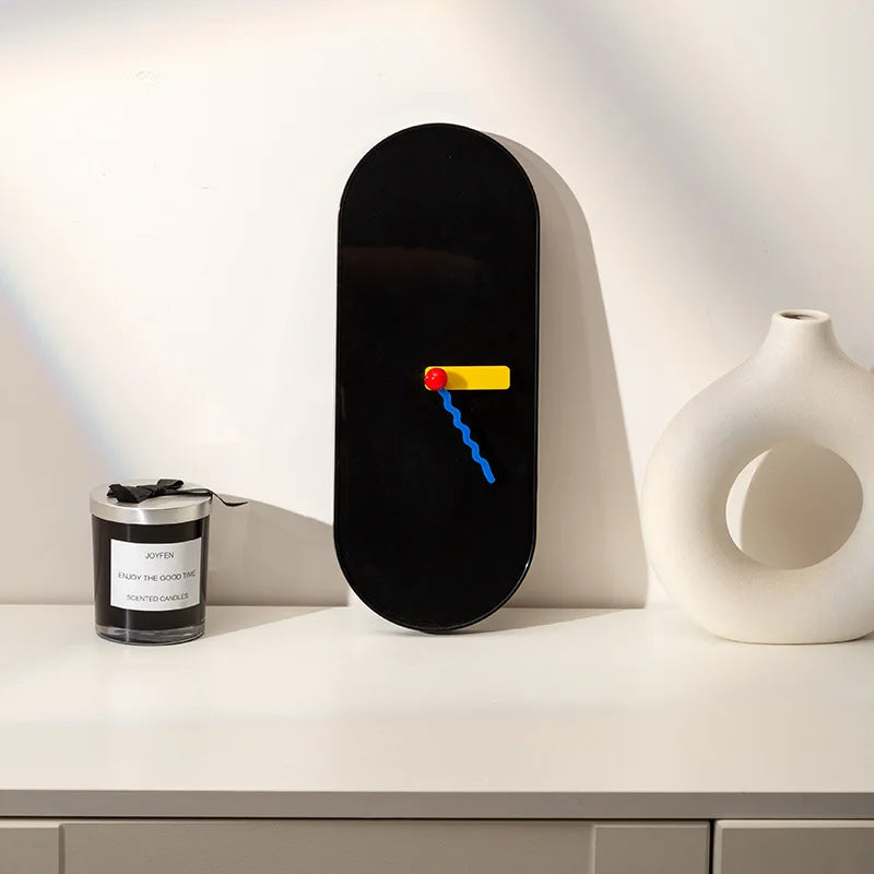 Black wall clock with colorful design on a white surface next to a candle and vase.